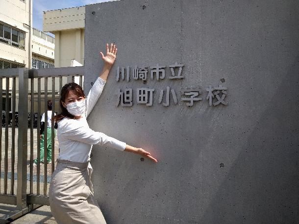 【写真】プレゼンターが川崎市立旭町小学校の前で写っています