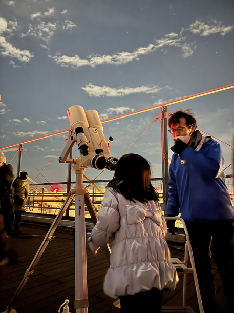 双眼鏡で月を観察している参加者にかわさき宙と緑の科学館の職員が解説している写真