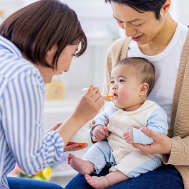 乳児が父親に前向きに抱きかかえられ、母親がスプーンで乳児に食事を与えている様子