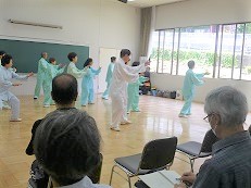 集会室でおそろいの衣装を着て太極拳の演舞する人々と観客